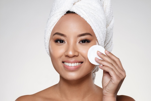 Pretty cheerful lady with towel on head cleaning her face