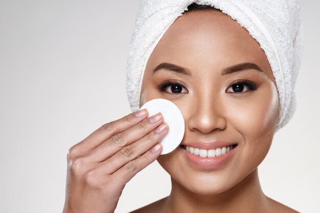 Pretty cheerful lady with towel on head cleaning her face