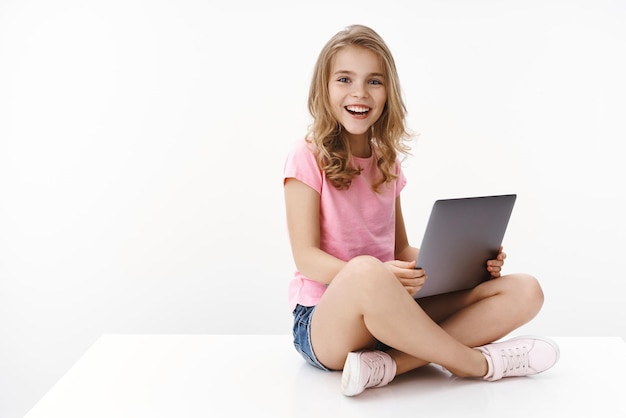 꽤 쾌활한 어린 소녀는 다리를 꼬고 앉아 집에서 노트북을 들고 흥분하며 인터넷 접촉 언어 교사 화상 통화 수업 흰색 배경을 통해 새로운 정보를 학습합니다.