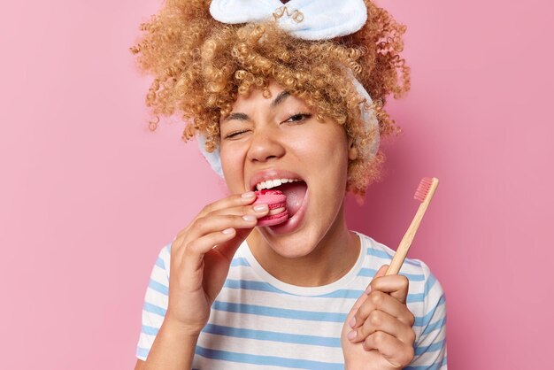 かなり陽気な女性はおいしいマカロンを食べますあなたの歯のために有害な食べ物を気にしません木製の歯ブラシは縞模様のTシャツを着ていますエナメル質のデザートの害