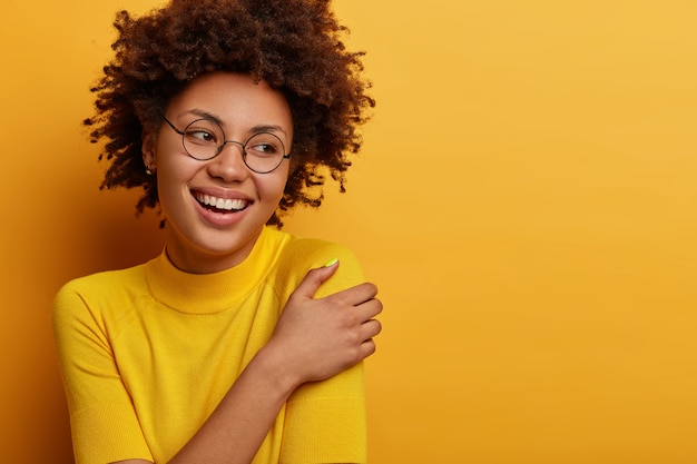 La donna abbastanza allegra guarda teneramente da parte, ha un sorriso felice, tocca delicatamente la spalla, ricorda piacevoli ricordi, ammira una splendida vista, ha i capelli ricci e folti, indossa gli occhiali, guarda pensierosa