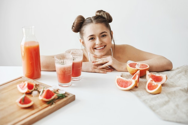 Foto gratuita donna abbastanza allegra con seduta sorridente dei panini alla tavola con le fette di frullato sano di dieta della disintossicazione del pompelmo sopra la parete bianca.