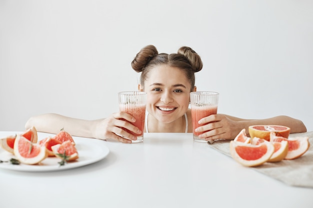 白い壁にグレープフルーツの健康的なデトックスダイエットスムージーのスライスをテーブルに座って笑顔のパンとかなり陽気な女性。