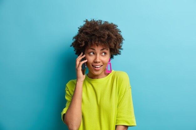 Pretty cheerful woman with curly hair has telephone conversation talks via mobile phone has glad expression