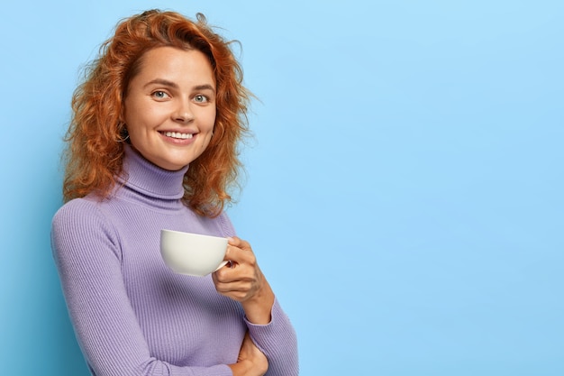 Pretty cheerful woman with ginger hair stands half turned to camera with white mug of coffee or tea