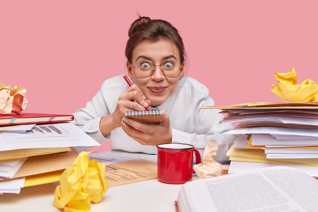 Pretty cheerful woman writes down list of things in notepad, holds pen, wears big optical glasses, has dark hair combed in knot, poses at workplace with documents