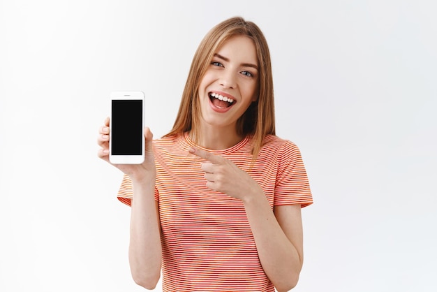 Бесплатное фото Довольно веселая молодая кавказская женщина в полосатой футболке держит смартфон, указывая на черный дисплей мобильного телефона, и улыбается, обсуждая новое приложение, рекомендует следить за ее белым фоном в социальных сетях