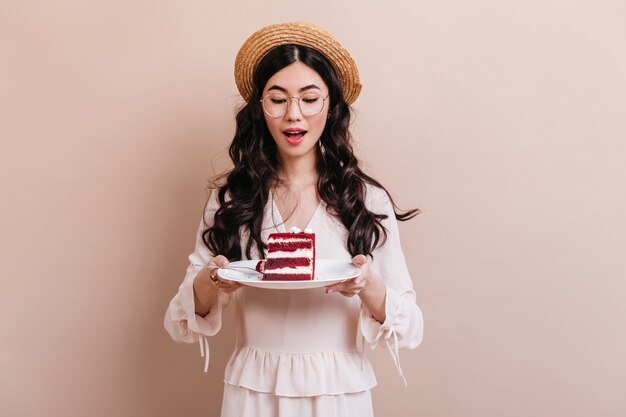 Pretty chinese woman in glasses holding plate with dessert. Attractive curly asian woman looking at cake.