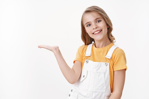 Pretty clever little girl, child with blond hairstyle in yellow t-shirt and overalls hold something on palm, introduce product blank white copy space, smiling joyful, brag what she got b-day present