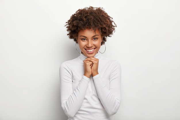 Pretty crisp girl with Afro hair, keeps both hands together under chin, has glad expression, wears round earrings, poloneck jumper