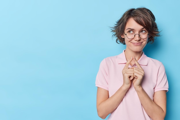 Free photo pretty cunning girl with bob hairstyle has idea makes plan schemes something smiles pleasantly dressed in casual t shirt and round spectacles isolated over blue background blank space for your promo