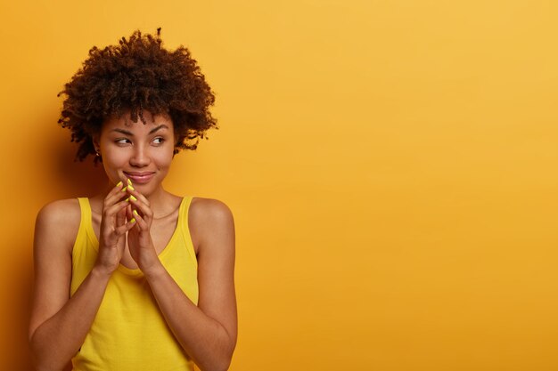 pretty cunning woman steepls fingers, looks mysteriously aside, has evil plan, wants to prepare surprise for husband, wears casual shirt, isolated on yellow wall, copy space area