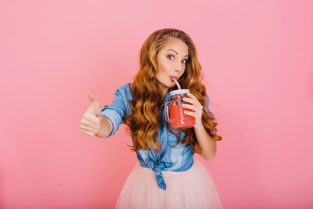 Pretty curly girl in trendy denim shirt shows a super sign with thumb up while trying fresh fruit smoothie. Portrait of adorable long-haired young woman in lush skirt drinking cocktail with pleasure