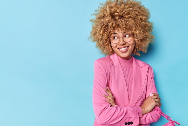 Pretty curly haired woman dressed in elegant pink jacket keeps arms folded smiles pleasantly holds bag focused away isolated over blue background with copy space for your promotional content