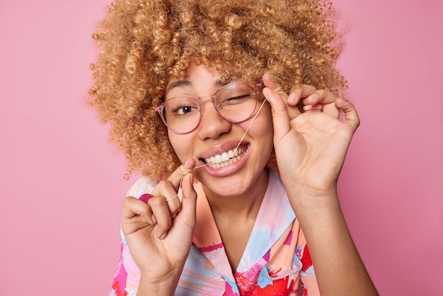 かなり縮れ毛の若い女性がデンタルフロスで歯をきれいにし、食べ物のウィンクを取り除きます目は光学ガラスを着用し、ピンクの背景に対してカラフルなシャツのポーズ口腔衛生と歯のケアの概念