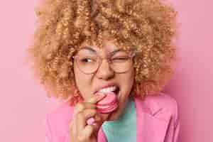 Free photo pretty curly woman bites hungrliy sweet macaroon winks eyes looks somewhere has addiction to sugar wears spectacles and formal clothes isolated over pink background delicious confectionary