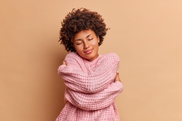 pretty curly woman embraces herself and closes eyes feels comfort in soft warm knitted sweater enjoys home tenderness tilts head poses against beige wall