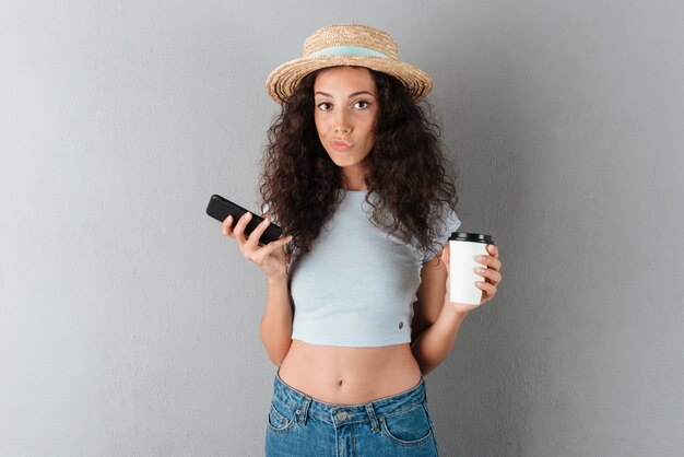 Довольно кудрявая женщина в шляпе с чашкой кофе и смартфон, глядя на камеру на сером фоне