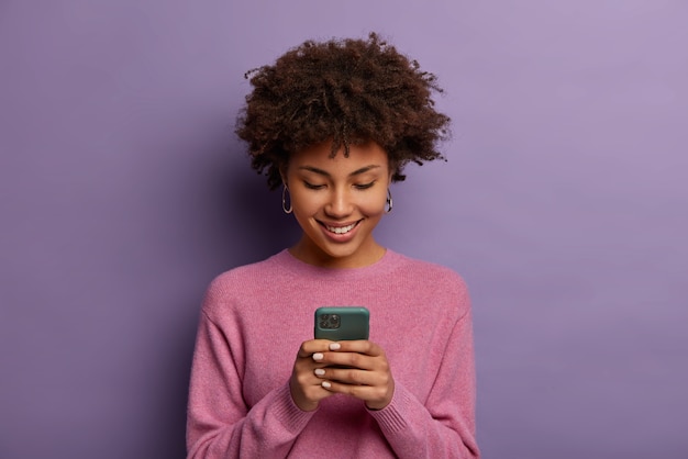 Pretty curly woman holds modern mobile phone, types messages on smartphone device, enjoys online communication, downloads special app for chatting, smiles tenderly, isolated on purple  wall