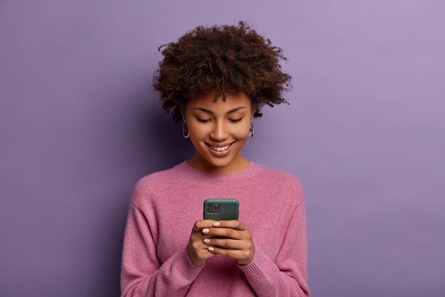 Симпатичная кудрявая женщина держит современный мобильный телефон, набирает сообщения на смартфоне, наслаждается онлайн-общением, загружает специальное приложение для чата, нежно улыбается, изолирована на фиолетовой стене