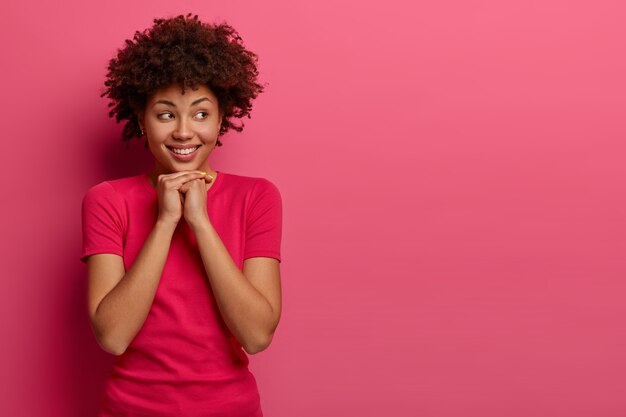 Pretty curly woman keeps hands under chin, chuckles positively and looks on right side, fascinated by something appealing, wears casual t-shirt, isolated on pink wall, free space aside