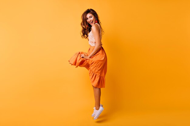 Красивая кудрявая женщина в оранжевой юбке прыгает с улыбкой Удивительная европейская дама в летнем наряде позирует перед желтой стеной