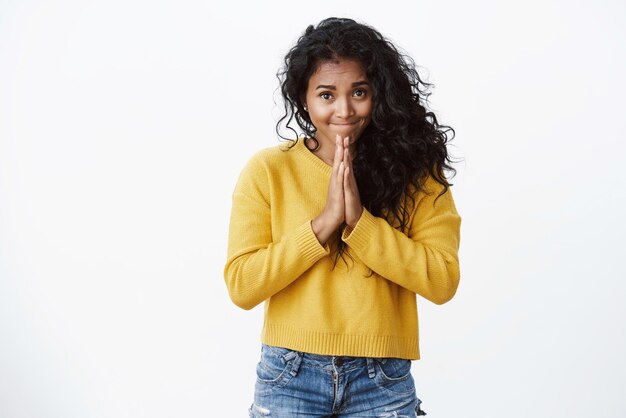 黄色いセーターを着たかわいいアフリカ系アメリカ人の縮れ毛の女性は、唇をすぼめ、助けやアドバイスを求めて物乞いをしているように愚かな笑みを浮かべて、手を一緒に押してお金を貸します