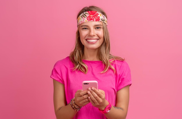 Pretty cute smiling woman in pink shirt boho hippie style accessories smiling emotional fun posing on pink background isolated positive mood using smartphone