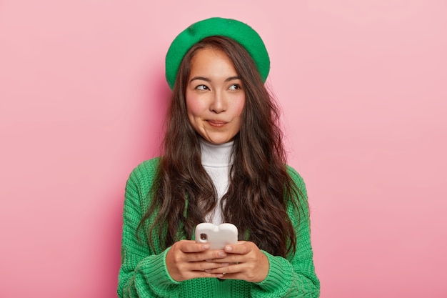 Pretty dark haired Japanese woman uses modern cell phone for sending text messages, surfes internet, has thoughtful expression