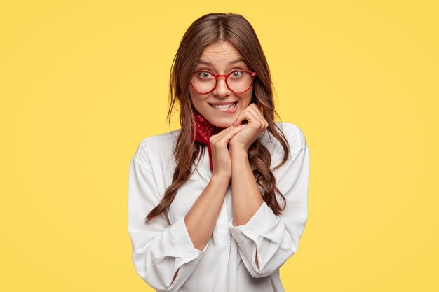 Pretty dark haired woman feels temptation, bites lower lip, looks with great desire, keeps hands near face, wears oversized white shirt, models against yellow wall, has red rimed spectacles