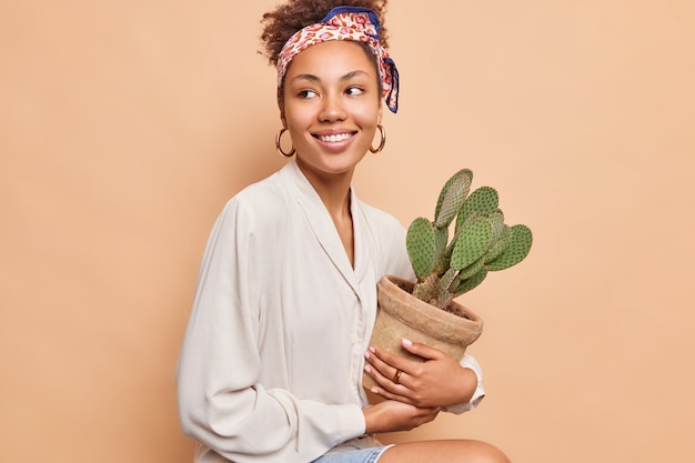 Pretty dark skinned curly haired woman sits looks happily away holds pot ot prickly succulent cactus