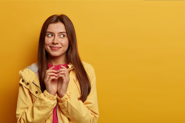 Pretty delighted woman schemes something, steepls fingers and looks with intention aside, has cunning look, made up good idea, smiles pleasantly, wears windbreaker, isolated on yellow  wall