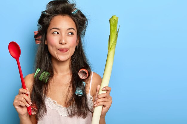 Pretty dreamy woman licks lips, holds spoon and fresh vegetable, looks aside, ready for preparing delicious nutritious dish, models indoor