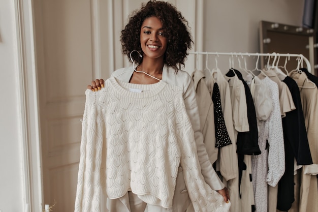 Free photo pretty elegant dark-skinned woman in beige jacket smiles, looks at front and holds hanger with white knitted sweater