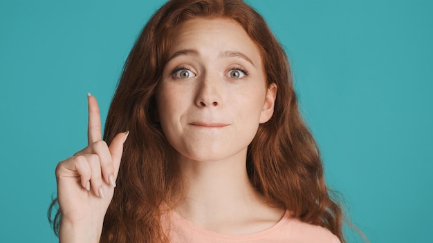 Foto gratuita ragazza rossa piuttosto emotiva che tiene il dito in alto guardando eccitato nella fotocamera che mostra un nuovo gesto di idea su sfondo colorato