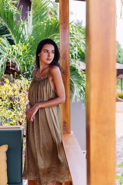 Pretty european positive smiling woman in summer flying dress at natural day light at villa terrace enjoying beautiful vacation, outdoor sofa with pillows on tropical