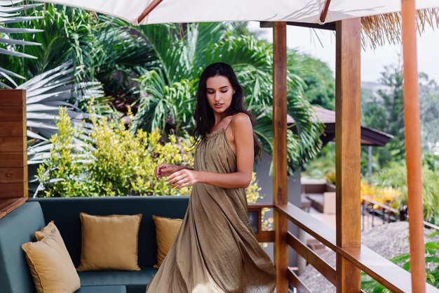 Pretty european positive smiling woman in summer flying dress at natural day light at villa terrace enjoying beautiful vacation, outdoor sofa with pillows on tropical