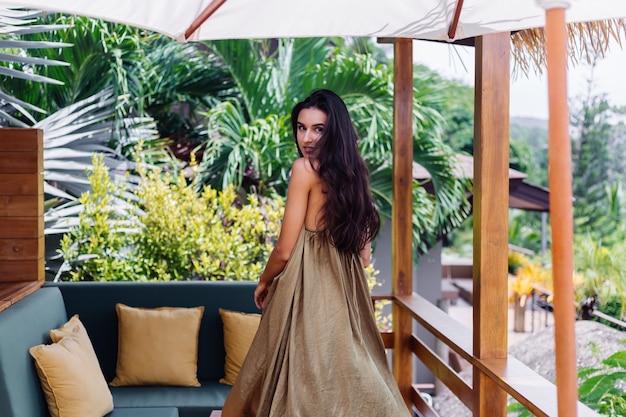 Pretty european positive smiling woman in summer flying dress at natural day light at villa terrace enjoying beautiful vacation, outdoor sofa with pillows on tropical