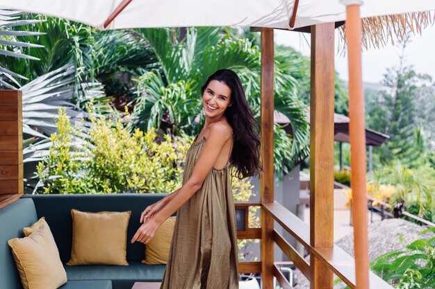 Pretty european positive smiling woman in summer flying dress at natural day light at villa terrace enjoying beautiful vacation, outdoor sofa with pillows on tropical.