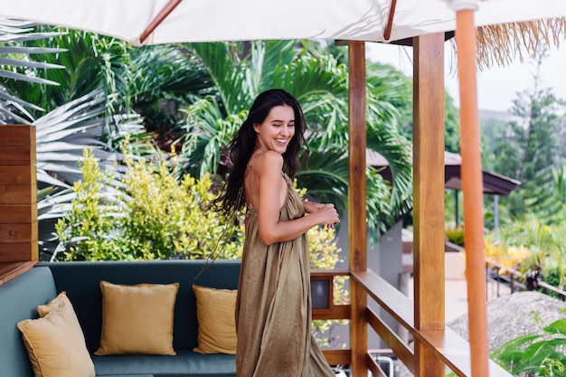 Pretty european positive smiling woman in summer flying dress at natural day light at villa terrace enjoying beautiful vacation, outdoor sofa with pillows on tropical.