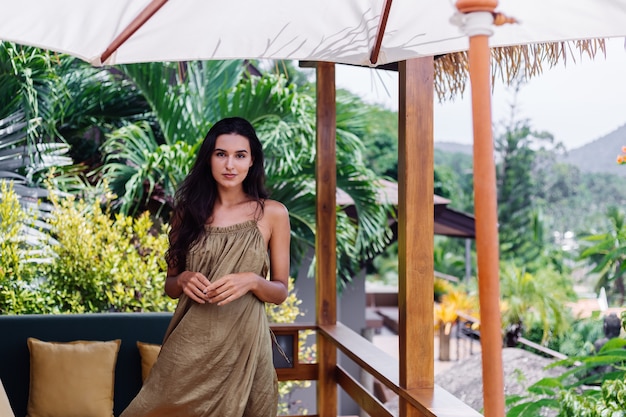 Pretty european positive smiling woman in summer flying dress at natural day light at villa terrace enjoying beautiful vacation, outdoor sofa with pillows on tropical