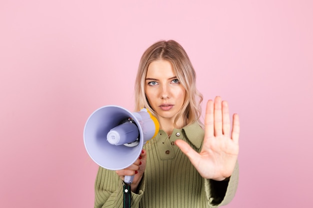 Pretty european woman in casual sweater on pink wall. unhappy serious with megaphone doing stop sign with palm of the hand