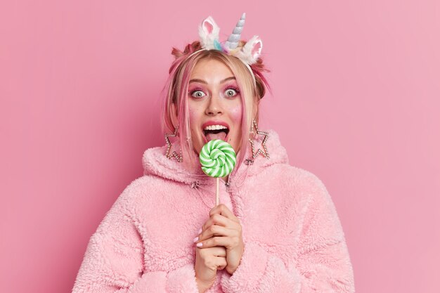 Pretty European woman has makeup licks delicious candy looks surprisingly at camera wears fashionable coat and unicorn headband 