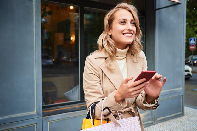 Pretty excited blond girl in stylish trench coat with shopping bags happily using cellphone on city street