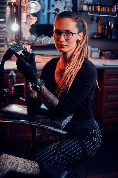 眼鏡をかけたかなり経験豊富なタトゥーアーティストが、タトゥーセッションの前に新しいタトゥーマシンを調整しています。