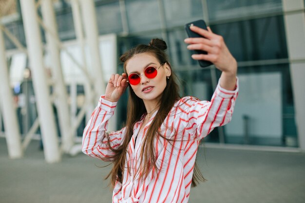 Pretty fashion model in red glasses takes selfie on her smartphone 