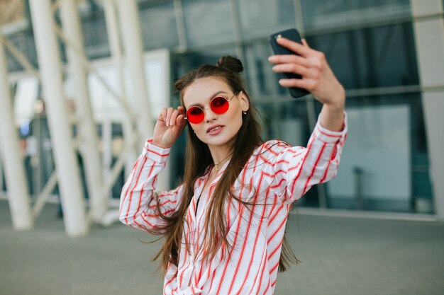 赤い眼鏡のかわいいファッションモデルは彼女のスマートフォンでセルフを取る