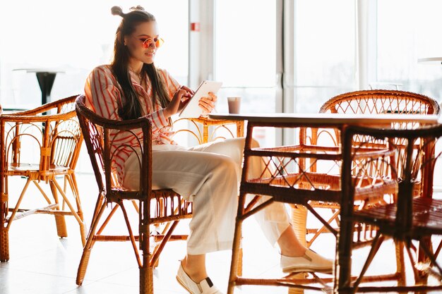 Довольно модель в красных солнцезащитных очках работает со своим планшетом, сидящим в кафе