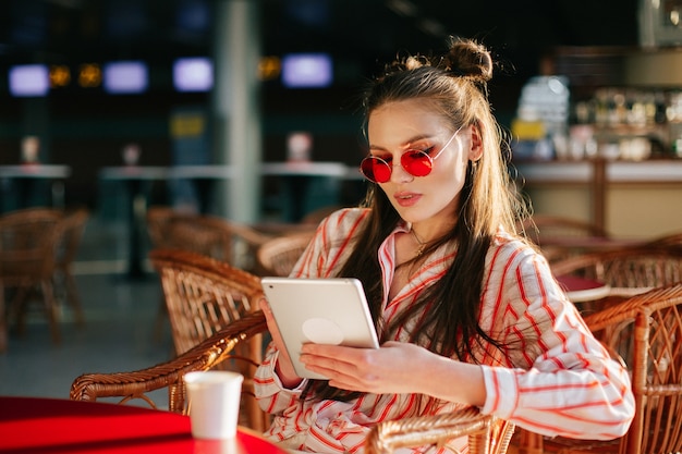 赤いサングラスのかわいいファッションモデルは、カフェに座っている彼女のタブレットで動作します