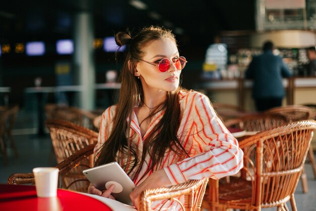 빨간 선글라스에 예쁜 패션 모델은 카페에 앉아 그녀의 태블릿과 함께 작동